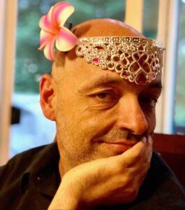 Jeremy White wearing an upside-down tiara with an orchid over his right ear, chin resting in his left hand