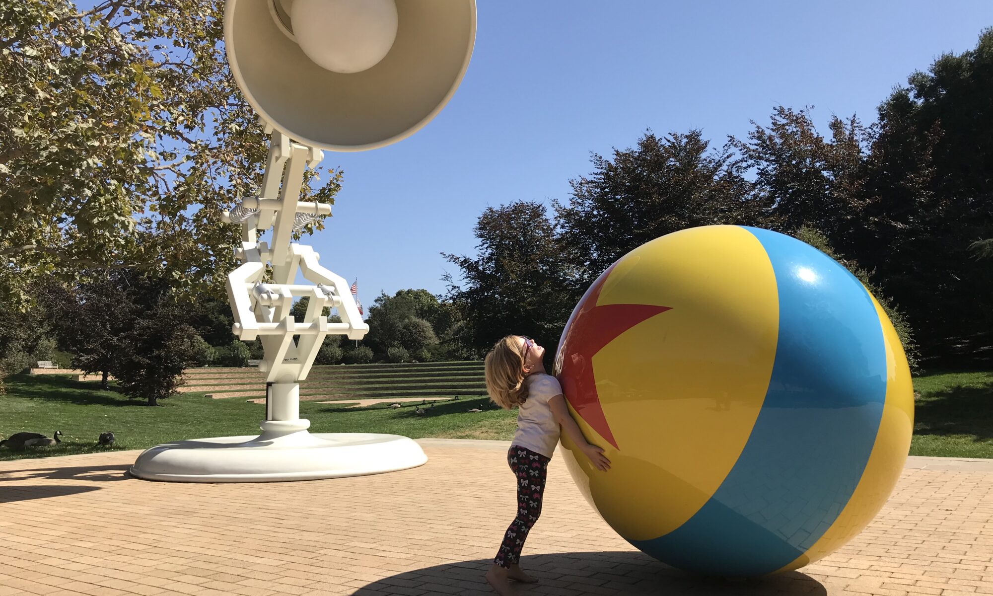 A young girl attempts to move a large model of the Pixar ball in a bricked courtyard overseen by it's companion Luxo.